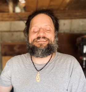 A man with a beard and mustache smiles with his eyes closed. He is wearing a light gray t-shirt and a necklace with a pendant. The background appears to be an indoor space with wooden elements.